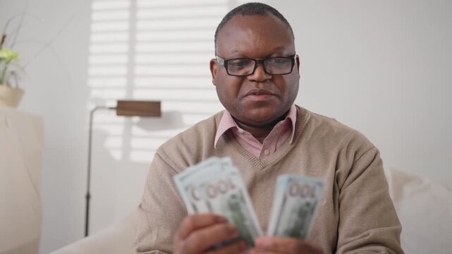 Happy middle-aged african american man counting new hundred dollars bills. Medium portrait of black male person in home interior, salary and fee for good job, successful financial deal of businessman