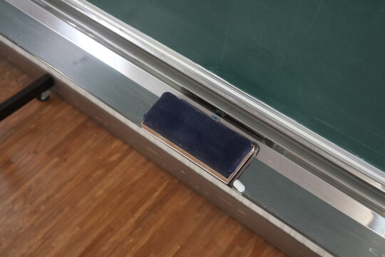 Green chalkboard with metal frame and dark eraser. Eraser rests beside polished silver rail. Floor shows warm wooden texture beneath. Still scene suggests quiet classroom or study space