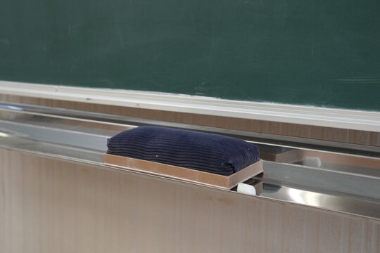 Dark eraser rests on metal ledge. Chalkboard green surface looms above. Quiet classroom stillness holds the scene. Ideal for education, teaching, or learning themes. No people, no animals