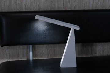 Minimalist desk lamp casts soft light. Modern design stands on dark surface. Backrest of chair provides contrast. Light emphasizes clean, contemporary style. Ideal for interior, design © Yoshida