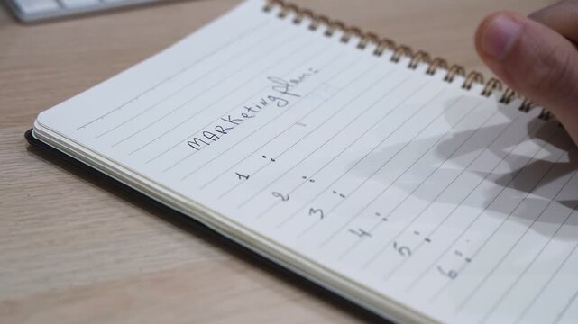 Close-up of a hand writing a marketing plan on a spiral notepad at a wooden desk, ideal for business, corporate strategy, and advertising content.