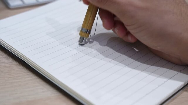 Close-up of a hand holding a wooden bamboo pen over a blank lined notebook on a light wooden desk, ideal for business, education, and corporate content.