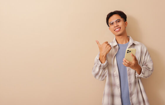 Excited young asian man holding mobile phone and pointing thumbs sideways while smiling, isolated on brown background in studio, expressing approval, promotion, or recommendation concept.