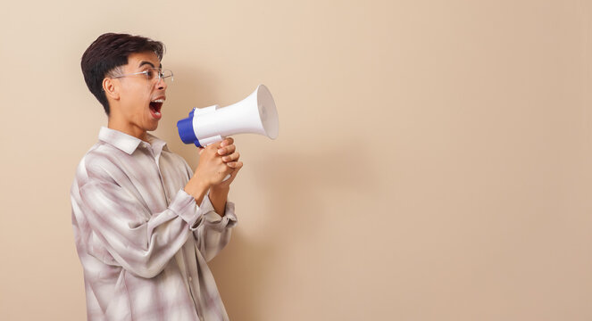 Excited young asian man shouting with a megaphonein a brown isolated background with copy space.