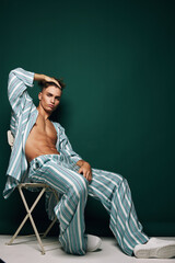 Young man in striped pajamas sitting on chair with hand on head against dark green background,...
