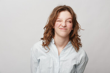 Obraz premium Young woman with curly hair wearing white shirt makes funny face with eyes closed and scrunched nose. Studio portrait on light background. Concept of humor, emotion, and casual style.