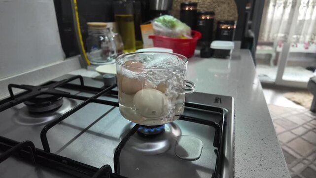 Three eggs boiling in a transparent glass pot on a gas burner, kitchen interior blurred background.