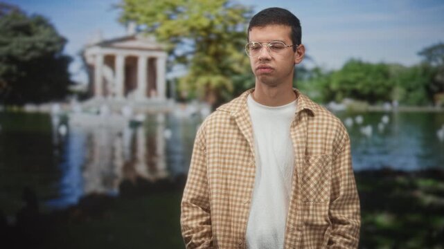 Young man with glasses stands with hands in pockets by a classical temple pond in park; contemplation.