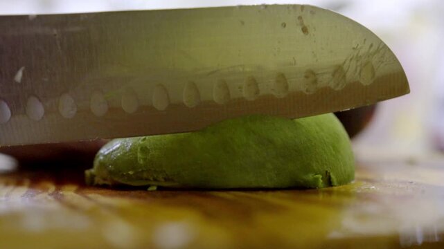 A sharp kitchen knife carefully slices through a fresh, green avocado on a rustic wooden board.