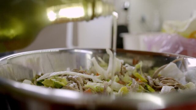 Close up of a chef pouring vegetable oil or dressing into a large stainless steel bowl filled with salpicon salad, featuring shredded meat and fresh vegetables.