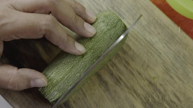 A person slices a green zucchini into long strips on a rustic wooden board.