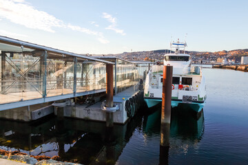 Fototapeta premium Trondheim fast boat terminal coastal view. A modern ferry moored at a glass-sided terminal along a calm waterfront, city skyline and hills in the background. Norway