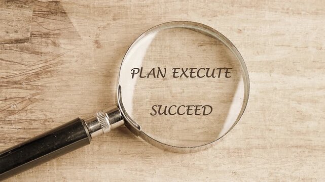 A close-up view shows a magnifying glass moving over a light wooden surface, gradually clarifying faint marks into the motivational phrase PLAN EXECUTE SUCCEED