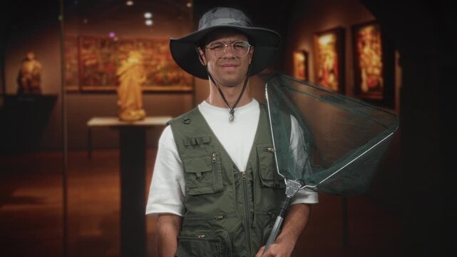 Man holds large fishing net over his shoulder and wears fishing vest and wide hat in museum building near sculpture and framed paintings; curiosity irony.