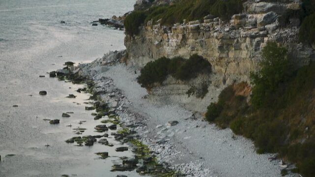 Drone tilt up from rocky shoreline to cliffs and lighthouse on Stora Karls&ouml; island at dawn with calm Baltic Sea and soft morning light, Gotland Sweden