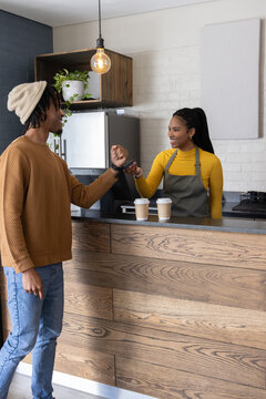 African American customer and barista in gray apron fist bumping at cafe counter with paper cups