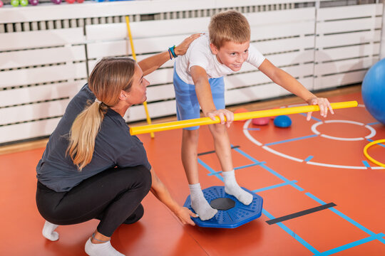 Motoric balance exercise on a stability board while holding a stick for posture control under trainer supervision.