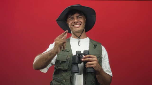 Man in hat and field vest holding binoculars and making rock horns gesture in studio with red backdrop; playful exploration.