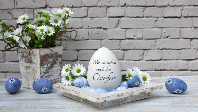 Gro&szlig;es wei&szlig;es Osterei mit der Aufschrift Wir w&uuml;nschen ein frohes Osterfest mit wei&szlig;en G&auml;nsebl&uuml;mchen und blauen Ostereiern vor einer hellen Ziegelwand.