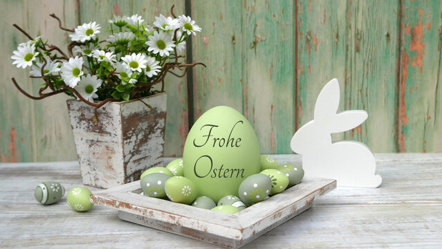 Gro&szlig;es gr&uuml;nes Osterei mit der Aufschrift 'Frohe Ostern' Ostereiern auf einem Holztablett neben einem wei&szlig;en Osterhasen aus Holz und einem Topf mit wei&szlig;en G&auml;nsebl&uuml;mchen vor gr&uuml;nlich verwittertem Holz