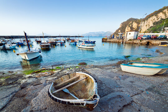 Small fishing boats moored in calm water of Marina Grande harbor on Capri island, Italy. Warm sunset light reflects on surface, illuminating coastline, pier, and distant mountains under clear sky.