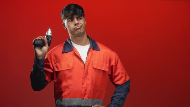 Man in orange work uniform holds a cordless drill aloft with one hand on hip and pouting expression in studio; bemusement manual work.