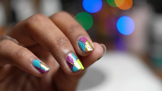 Close-up of a person's hands featuring geometric, colorful nail art with gold foil accents, using an electric nail file with a colorful bit attachment against a bokeh background.