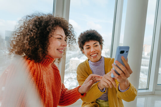 Smiling friends with curly hair talking and using phone by window
