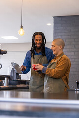 Diverse coworkers smiling and making coffee at cafe counter, wearing aprons, handling red cup © wavebreak3