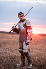 Fototapeta premium A medieval knight with a bloody face, dressed in plate armor with a sword and a torch in his hands, poses standing in a field against the backdrop of the sky and sunset.