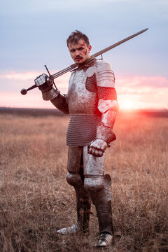 A medieval knight with a bloody face, dressed in plate armor with a sword and a torch in his hands, poses standing in a field against the backdrop of the sky and sunset.