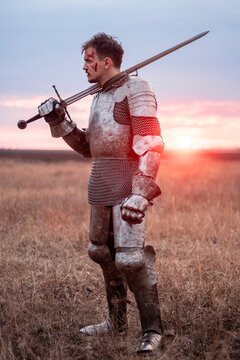 A medieval knight with a bloody face, dressed in plate armor with a sword and a torch in his hands, poses standing in a field against the backdrop of the sky and sunset.