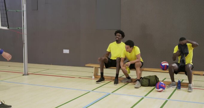 Diverse male volleyball team in yellow shirts during break walking to gym bench grabbing phone