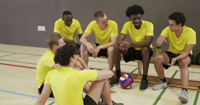 Diverse male teammates in yellow tops laughing, planning during break at gym with volleyball