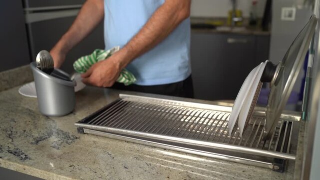 Man doing chores, wiping a wet plate with a tea towel over a drying rack in the kitchen