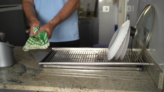 Male hands drying a spoon with a dishcloth and placing it on a metal rack in the kitchen