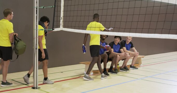 Diverse yellow-shirt men entering left, walking gym court with duffel greeting blue team