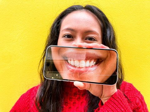 Woman holding phone with artificial smile in front of face against yellow wall