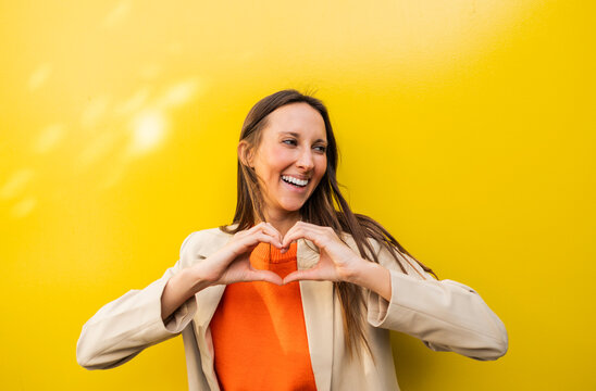 Smiling woman making heart gesture against bright yellow wall