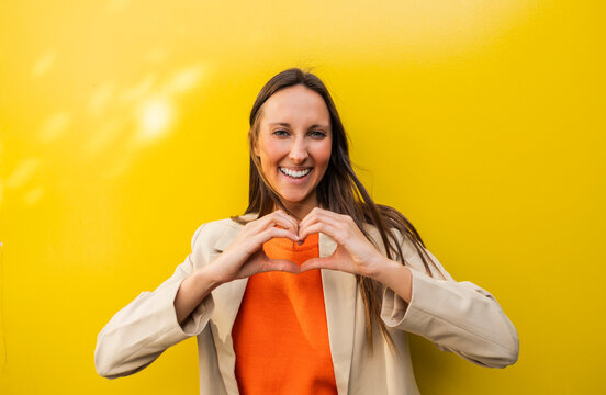 Smiling woman making heart gesture in front of yellow wall