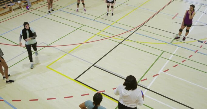 Women training with coach in white sweatshirt raising arm, tossing purple balls on gym court