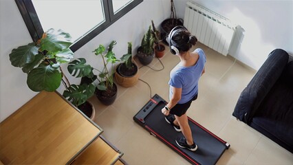 Woman exercising at home, walking on a treadmill while listening to music with headphones, focusing on personal fitness and wellness in an indoor environment © DawDunia