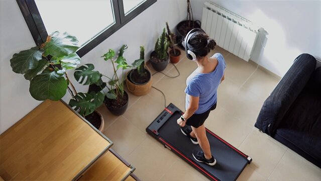 Woman exercising at home, walking on a treadmill while listening to music with headphones, focusing on personal fitness and wellness in an indoor environment