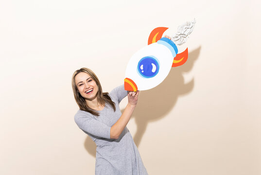 Smiling woman in gray dress holding handmade paper rocket indoors