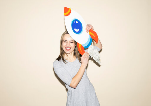 Smiling woman in gray dress holding handmade paper rocket indoors