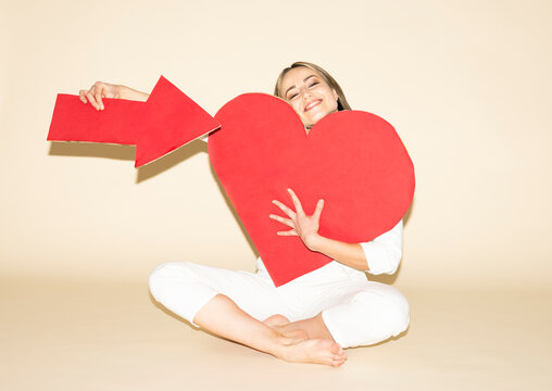 Woman in white jumpsuit holding red heart and arrow symbol indoors