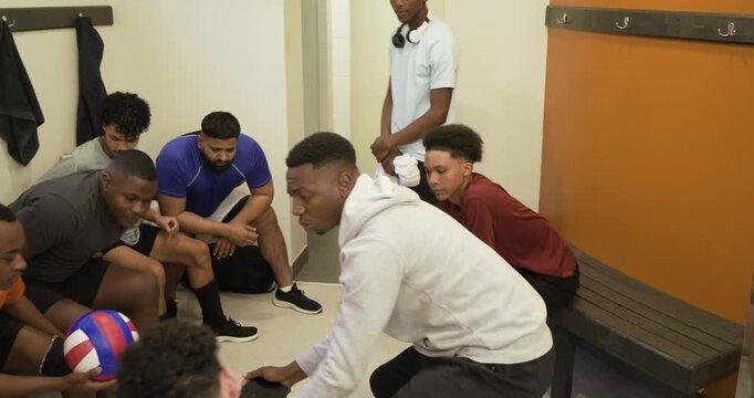 Male team gathering in locker room while hooded captain starting pre-game pep talk, with volleyball