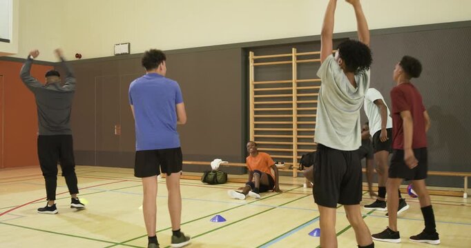 Diverse male team and coach following instruction, marching in school gym past cones and volleyball