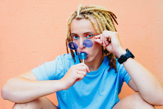 Portrait of young woman with dreadlocks, sunglasses and lollipop against orange wall