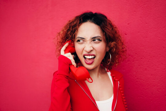 Redhead with curly hair making grimace while talking on red telephone
